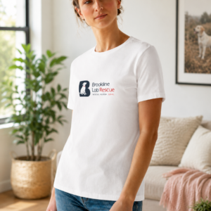 Woman wearing a white Brookline Lab Rescue T shirt with the rescue logo on the left chest, standing in a bright living room with modern décor and a framed Labrador Retriever photograph in the background.