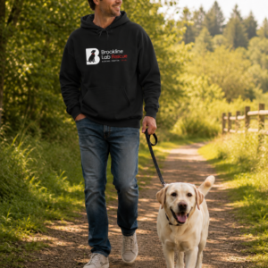 Man walking a yellow Labrador Retriever on a wooded trail while wearing a black Brookline Lab Rescue Gildan 18500 hoodie with the organization logo.