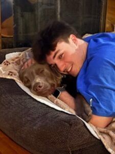 A young man smiles while cuddling a sleeping silver Labrador Retriever curled up in a dog bed.