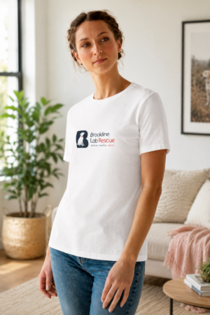 Woman wearing a white Brookline Lab Rescue T shirt with the rescue logo on the left chest, standing in a bright living room with modern décor and a framed Labrador Retriever photograph in the background.