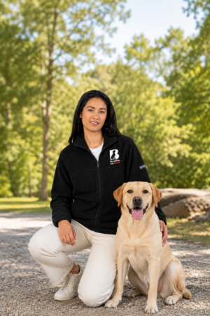 Woman wearing a black Brookline Lab Rescue fleece jacket beside a yellow Labrador outdoors